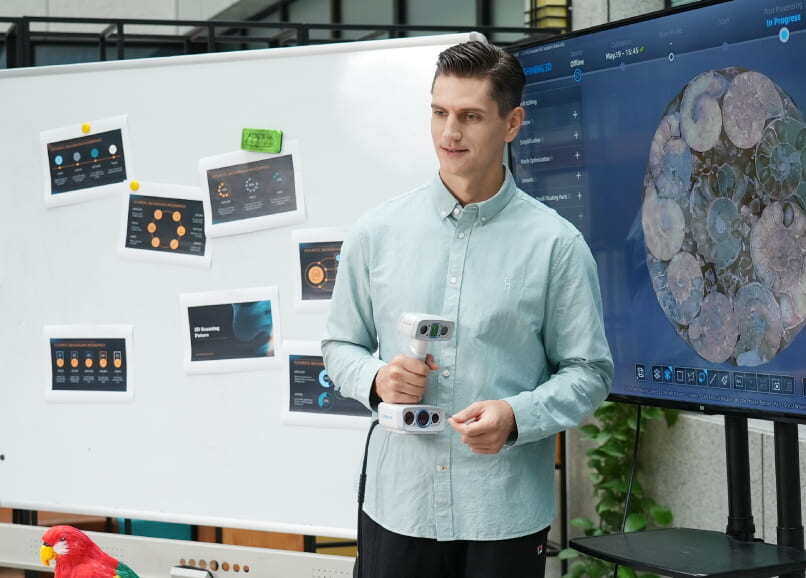Person holding the EinScan H2 scanner while presenting in front of a whiteboard and a large display screen.