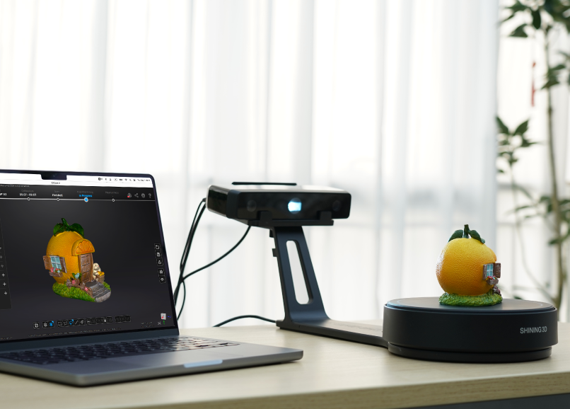 An EinScan SE V2 desktop 3D scanner capturing a small orange house model on the automated turntable, with the resulting 3D model displayed on a laptop screen.