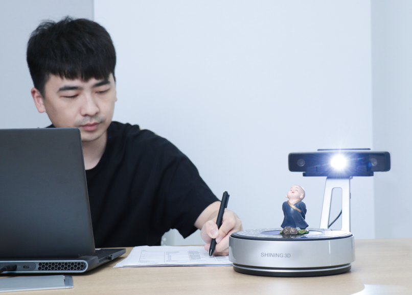 User working at a desk while the EinScan SP V2 scans a small monk figurine on the turntable.