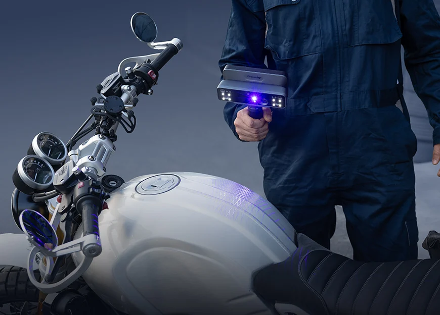 A technician scanning the fuel tank and handlebars of a motorcycle using the EinScan Rigil, with blue laser lines visible on the surface.
