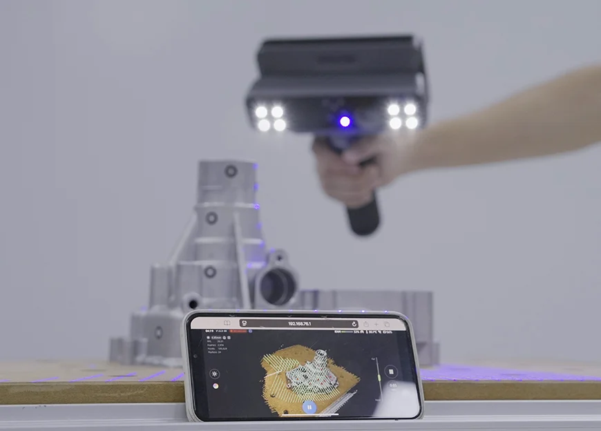 The EinScan Rigil scanning a metal mechanical part, while a mobile device on the table shows a live preview of the 3D scan.