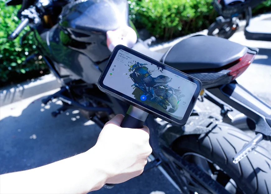 A person holding an EinScan Rigil scanner outdoors while capturing a 3D scan of a motorcycle, with the live scan preview displayed on the device’s screen.