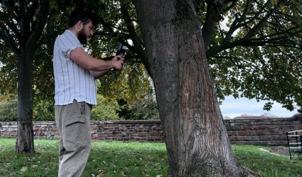 Designer using a handheld 3D scanner to capture the texture of a tree trunk outdoors.